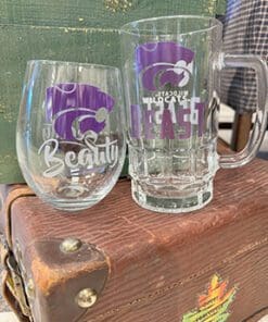 two glasses are displayed the one on the left is a wine glass with a K-State logo and beneath it reads "Beauty" and the one on the right is a beer mug displays the K-State logo and reads "Beast"